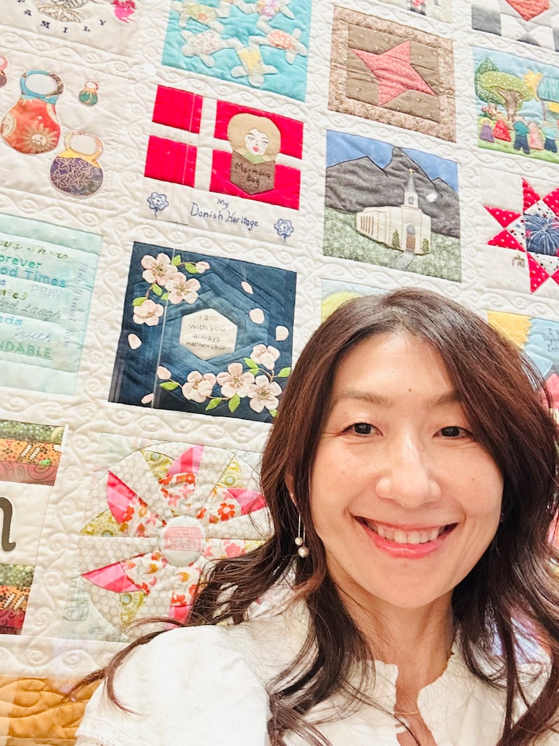 Sachiko Aldous stands by her quilt block, which is blue with cherry blossoms appliqued on top, in "The Family Quilt" display at the Temple Square Visitors' Center in Salt Lake City, July 2026.