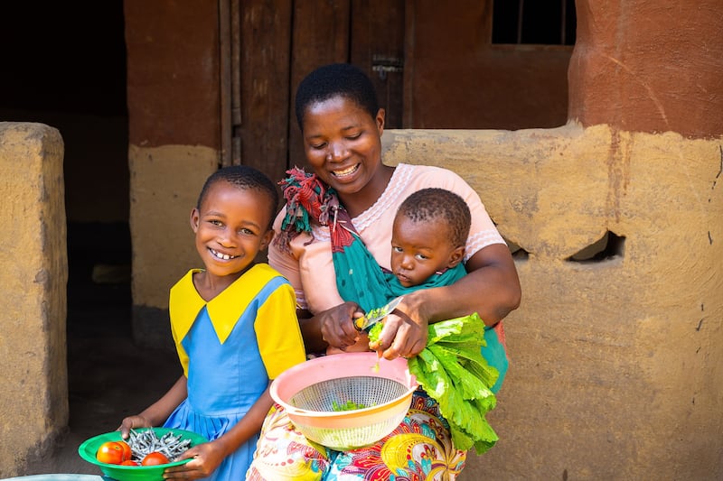 Une jeune fille est assise avec sa mère, qui porte son frère ou sa sœur de 1 an tout en coupant des légumes devant une maison dans le sud du Malawi.